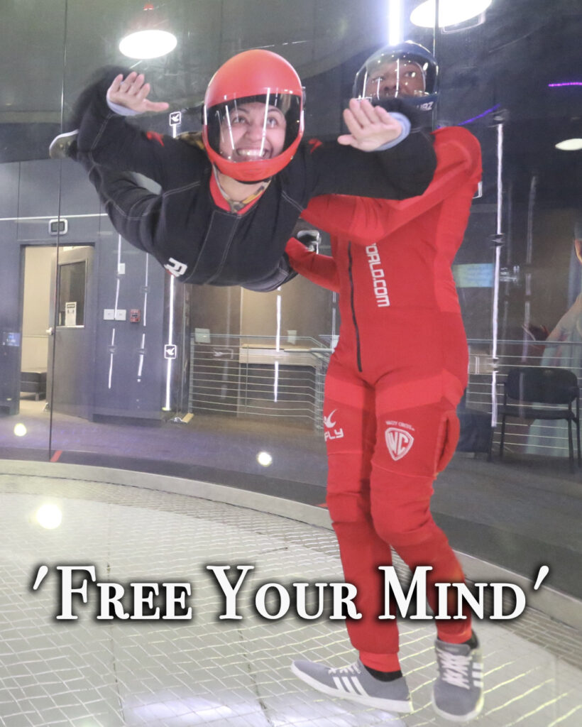 A man and woman in red suits are inside an indoor skydiving facility.
